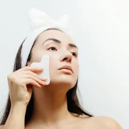 A woman using a white facial tool on her cheek, wearing a white headband in a bright, minimalistic setting.