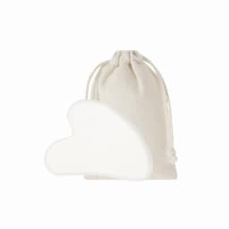 A heart-shaped white gua sha tool next to a natural fabric drawstring pouch.