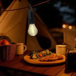 A cozy outdoor scene featuring a light bulb hanging above a table with a plate of food, cups, and a basket.