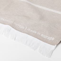 A light beige cotton towel with white fringe detailing and text woven into the fabric. It has a soft texture.