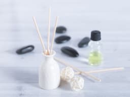 A white ceramic diffuser with wooden reeds, flanked by decorative balls and an essential oil bottle, on a light surface.