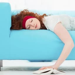 A woman with curly red hair rests on a light blue couch, with her head on the armrest and a book on the floor.