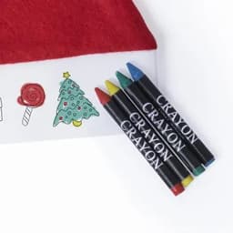 A set of four crayons in red, blue, green, and yellow, placed beside a decorated box with festive illustrations.