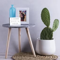 A table with a grey top and wooden legs, featuring a blue glass bottle, a picture frame, and a cactus in a white pot.