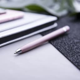 A pink pen with a silver tip rests on a textured surface, alongside a notebook and other stationery.