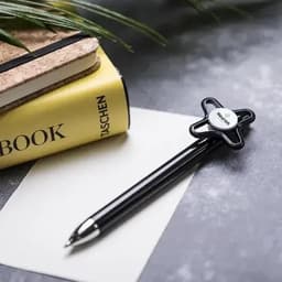 A black pen with a unique star-shaped grip rests on a grey surface alongside a yellow notebook and a piece of paper.