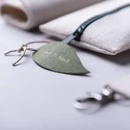 A green leaf-shaped tag is attached to a cream-coloured pouch with a zipper, featuring a small silver clasp.
