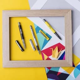 A collection of pens in black, silver, and yellow arranged within a wooden frame on a yellow geometric background.