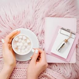 A drink bottle in silver is placed beside a pink notebook and a cup of coffee with marshmallows on a fluffy background.