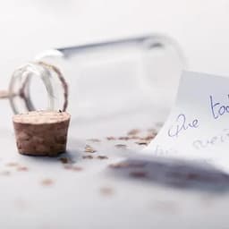 A small glass bottle with a cork on a surface scattered with confetti and a handwritten note beside it.