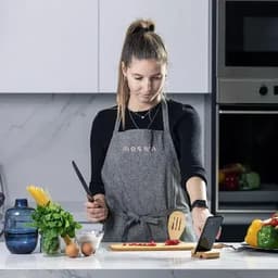 A woman in a grey apron prepares vegetables on a wooden board in a modern kitchen. A phone is positioned nearby.