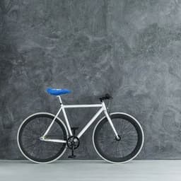 A bicycle with a white frame, black wheels, and a blue seat, positioned against a textured grey wall.