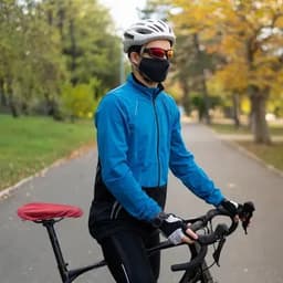 A cyclist in a blue and black jacket, wearing sunglasses and a mask, stands beside a bicycle in a park setting.