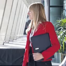 A woman in a red blazer holds a black folder, standing in a modern building with large windows and greenery.