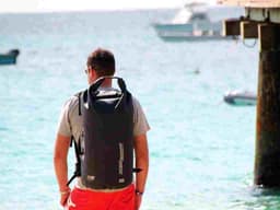 A black waterproof backpack is worn by a person facing the ocean, with boats visible in the background.