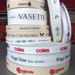 A stack of custom printed flat and herringbone cotton ribbon in cream and white featuring various logos.