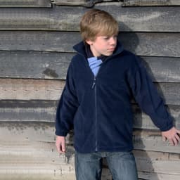 A youth in a navy polar fleece jumper stands against a wooden wall, featuring a full zip and a collar.