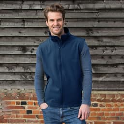 A navy softshell vest features a zip-up front and is styled with long sleeves. The model stands against a rustic wooden background.
