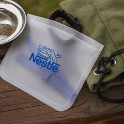A clear PEVA reusable food storage bag featuring a blue logo, placed on a wooden surface beside a plate and bag.