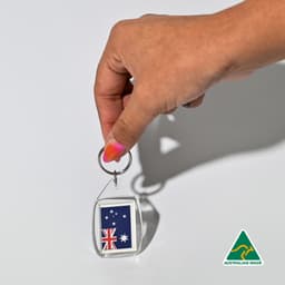 A hand holds a clear tear drop acrylic keychain featuring the Australian flag. The keychain has a silver ring and a logo.