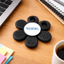 A black fidget spinner in a flower shape with a central logo, placed on a wooden desk near a coffee cup and stationery.