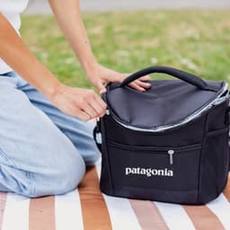 A black cooler bag with a zippered top and mesh pocket, sitting on a striped blanket. It has a logo.
