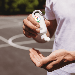 A white sunscreen bottle with a keychain cap, held above a hand, set against a blurred outdoor background.