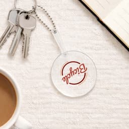 A clear PVC hand fan with a keychain, featuring a round shape, sits on a textured surface near a coffee cup and notebook.