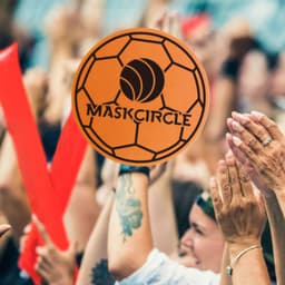 A crowd of people cheering, with hands raised and red foam mitts visible. An orange soccer ball logo is prominent in the centre.