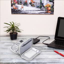 A grey electronic accessory organiser bag with multiple compartments, displayed on a desk alongside a laptop and a plant.