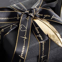 A black ribbon with gold edges, featuring a heartbeat design and text, tied around a dark gift box.