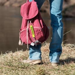 A keyring in yellow attached to a maroon backpack, held by a person standing on grass near a riverbank.