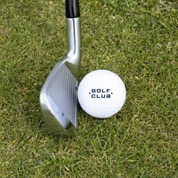 A golf ball in white positioned next to a golf club on green grass, featuring a logo on the ball.