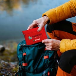 A bright red first aid kit pouch being taken from a green backpack near a lake. The pouch has a logo.