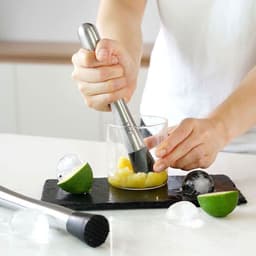 A stainless steel cocktail muddler is used to mash ingredients in a glass, set on a black slate surface with ice and lime.