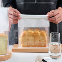 A person holds a transparent storage bag containing a loaf of bread, with a drink beside them. The bag has a white top.