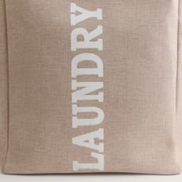 A laundry basket in a beige fabric with white lettering, featuring a sturdy handle and a simple design.