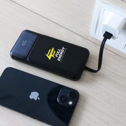 A black power bank with a logo is charging near a phone, connected to a wall socket with a cable.