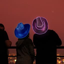 Light-up sequin cowboy hats in blue and pink, worn by two people against a city skyline at sunset.