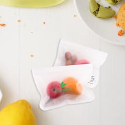 Two small transparent storage bags containing various colourful fruits, placed on a light wooden surface with some food items nearby.