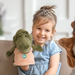 A green crocodile hand puppet held by a smiling child, wearing a light blue dress and a tiara.