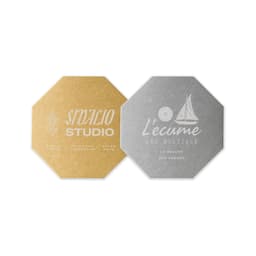 An octagonal metal coaster in gold and silver finishes, featuring logos on each side.