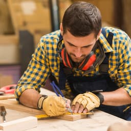 A man wearing a checkered shirt and gloves is measuring and marking wood in a workshop setting.