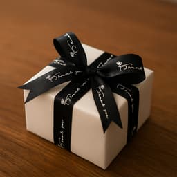 A cream gift box wrapped in black ribbon with a thank you message, featuring a bow on top.