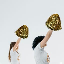 Gold cheerleading pom poms held up by two individuals wearing white t-shirts, with a simple background.