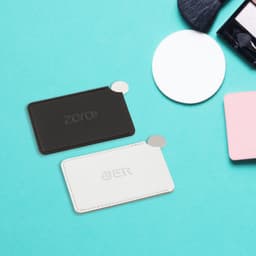 A black and white stainless steel portable mirror with a sleek design, placed on a light blue surface alongside beauty tools.