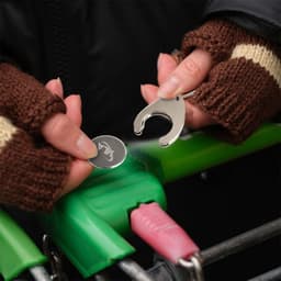 A silver teardrop-shaped trolley coin keyring is being placed into a shopping trolley, held by hands in brown knit gloves.