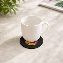 A white mug sitting on a black custom record coaster on a marble surface, beside a green plant and a magazine.