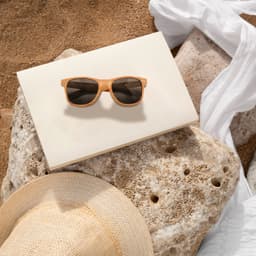 Wooden sunglasses with brown lenses resting on a light-coloured surface, surrounded by textured stones and a straw hat.