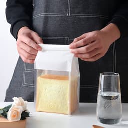 A clear bag with a white top seal containing a loaf of bread, positioned next to a glass of water.
